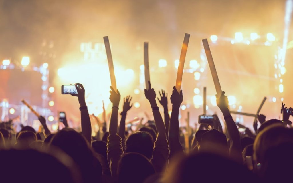 Rozentuzjazmowany tłum z uniesionymi rękami i świetlnymi pałeczkami podczas koncertu