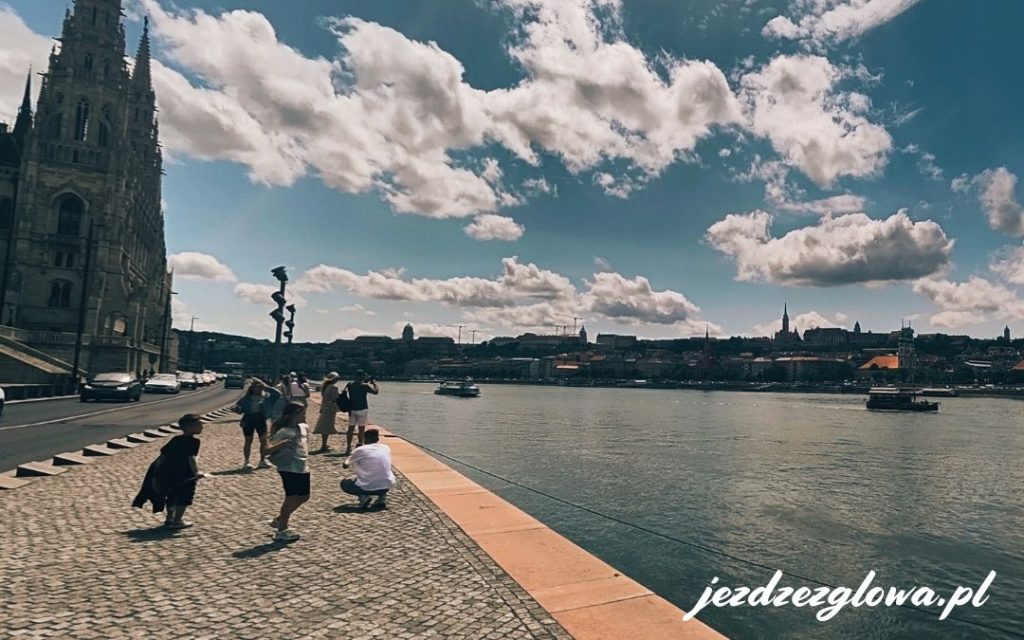 Widok na Dunaj w Budapeszcie obok Parlamentu, z turystami na nabrzeżu i panoramą Wzgórza Zamkowego po drugiej stronie rzeki