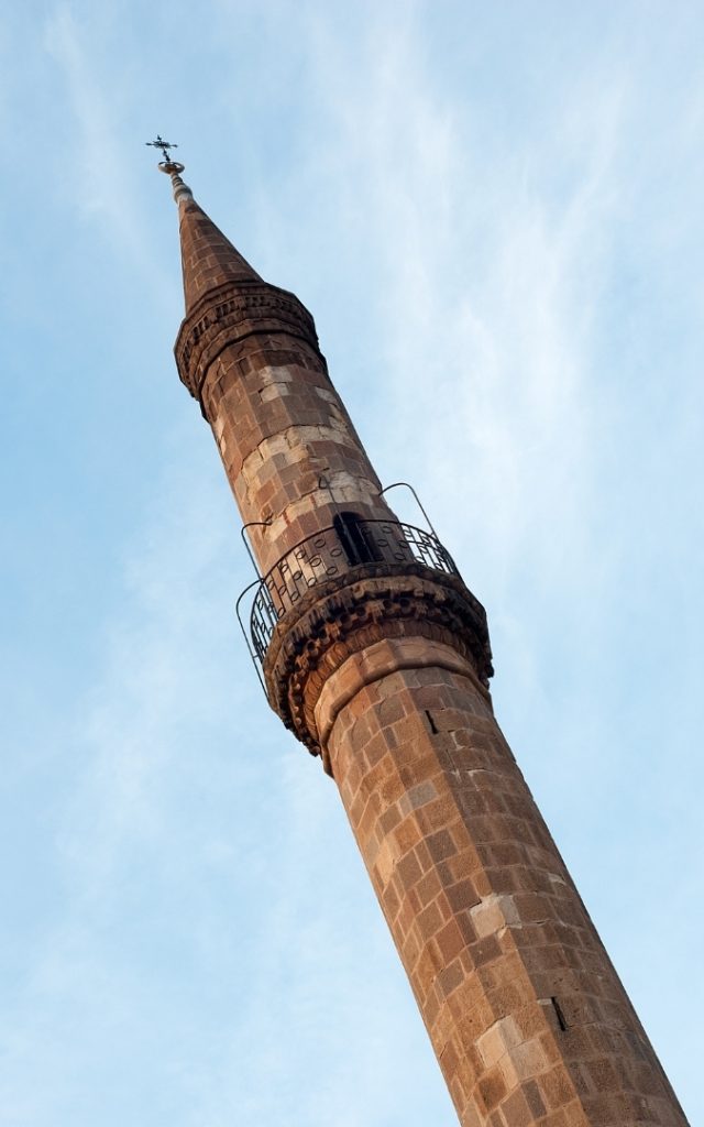 Ceglany minaret w Egerze, zwężający się ku górze, z balkonem otoczonym metalową balustradą, na tle jasnego nieba