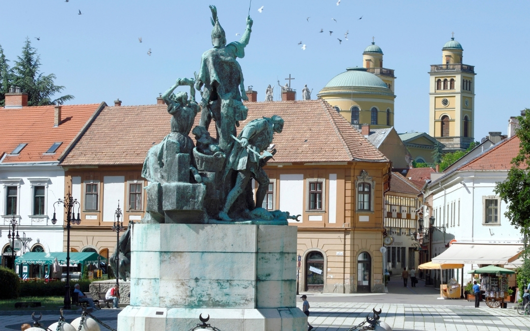 Pomnik Istvana Dobo w Egerze, przedstawiający bohaterów obrony zamku, na tle zabytkowych kamienic rynku, z widoczną w oddali Bazyliką w Egerze