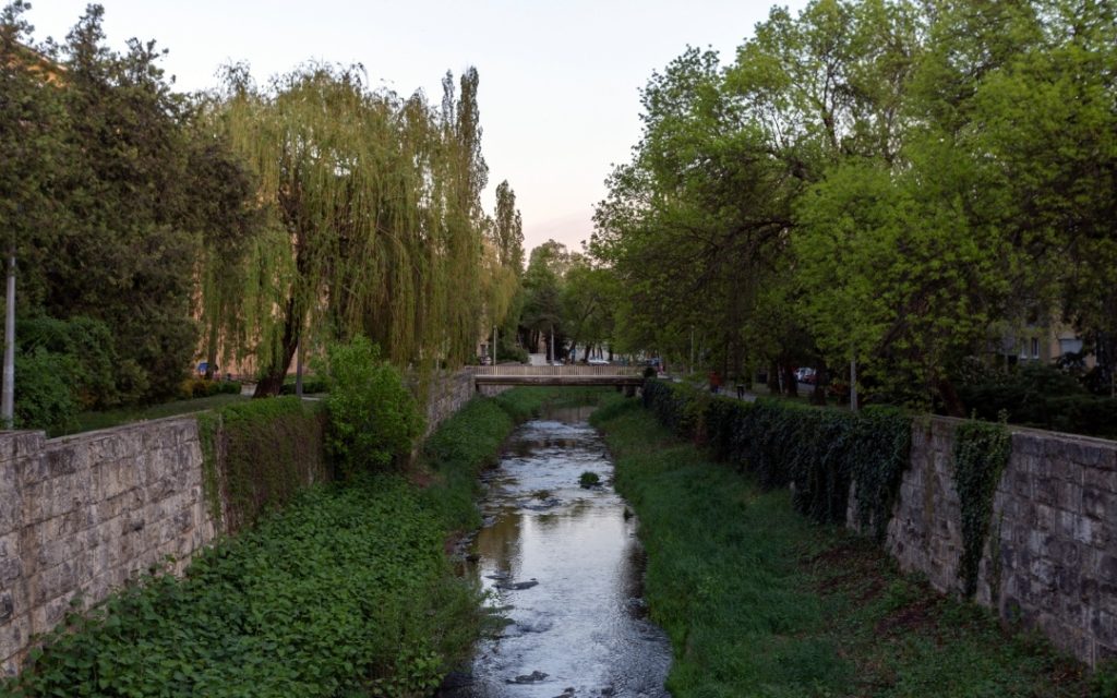 Strumień w Egerze otoczony kamiennymi murkami i zielenią, z mostkiem widocznym w tle