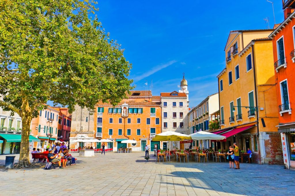 Plac Campo Santa Margherita otoczony pastelowymi budynkami, ogródkami kawiarnianymi i dużym cienistym drzewem