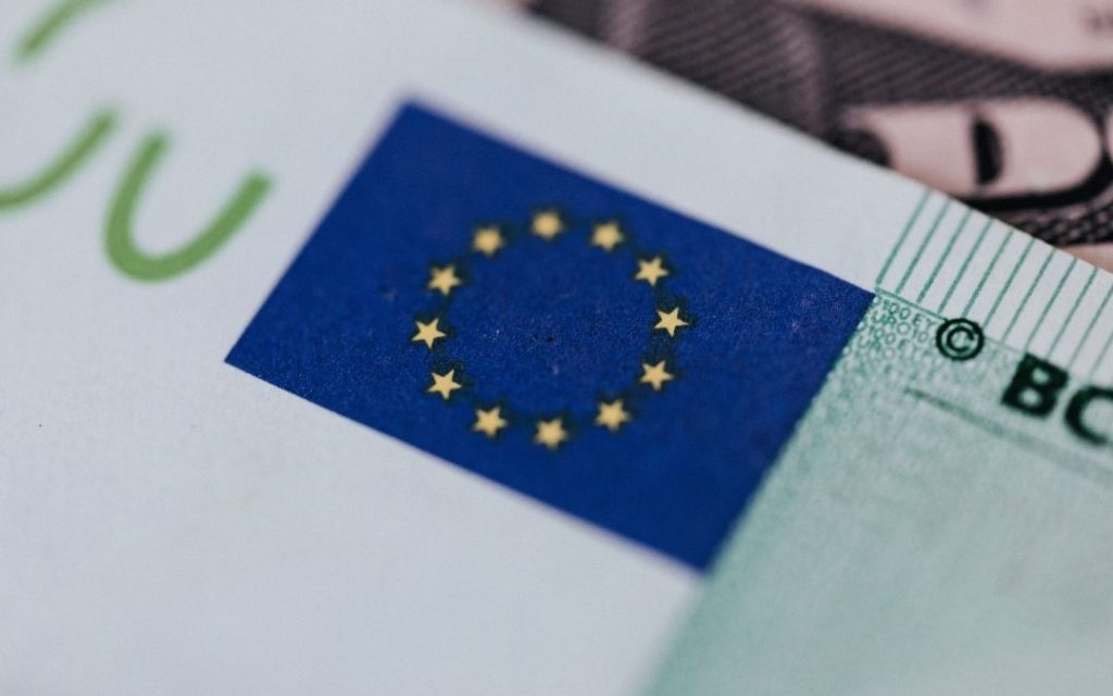 Bardzo bliskie ujęcie banknotu euro. W centrum ostrości znajduje się flaga Unii Europejskiej (niebieskie tło z dwunastoma gwiazdami).