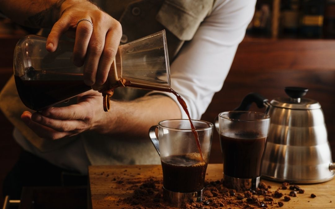 Barista nalewa kawę z szklanego naczynia (Chemex) do szklanej filiżanki. Na drewnianym blacie widać ziarna kawy i czajnik.