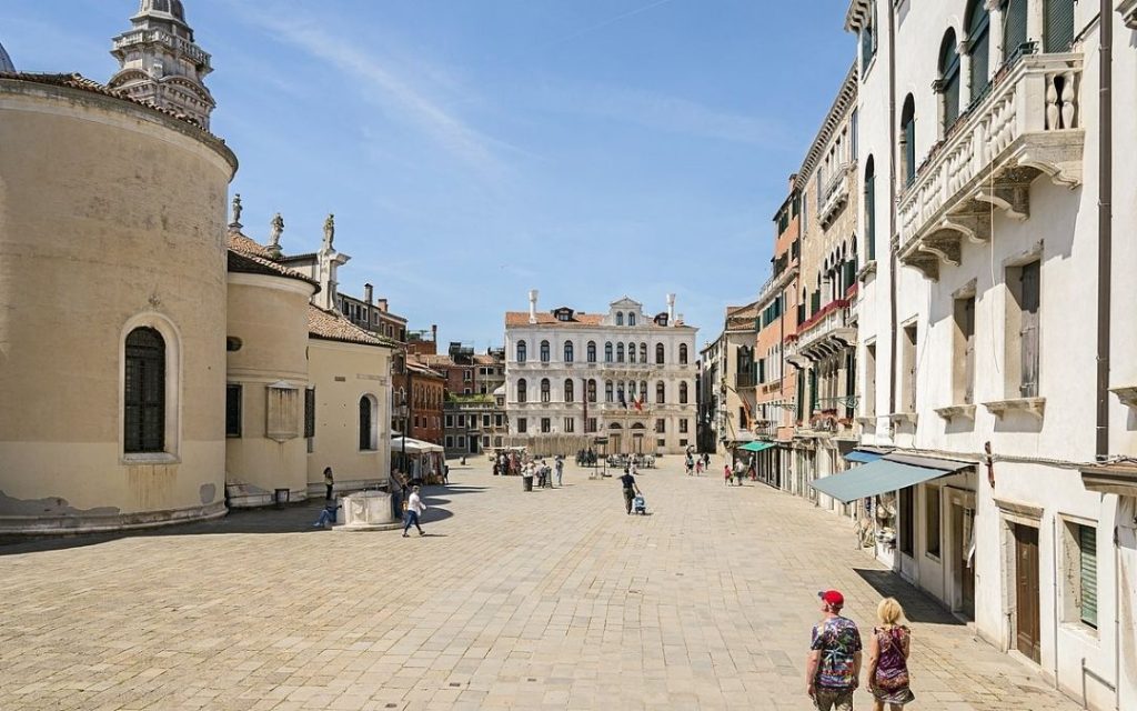 Campo Santa Maria Formosa w Wenecji. Słoneczny plac otoczony historycznymi kamienicami i Kościołem Santa Maria Formosa (widoczna apsyda) z przechodniami.
