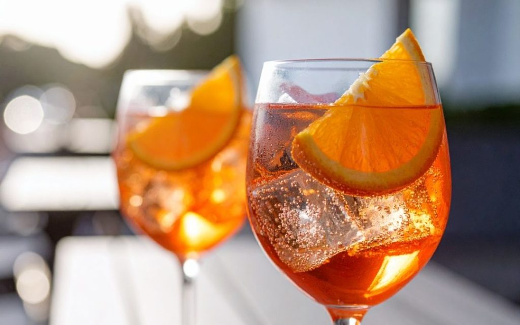 Dwa pomarańczowe drinki Aperol Spritz w kieliszkach. Napoje wypełnione lodem i ozdobione plastrami pomarańczy w pełnym słońcu.