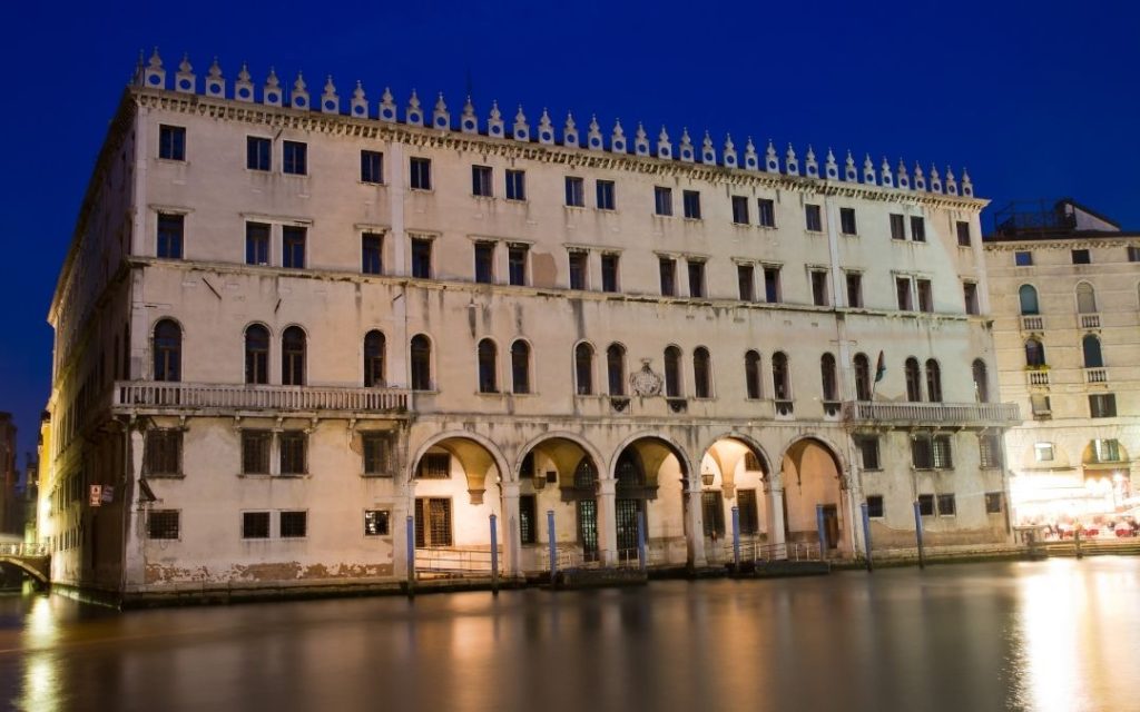 Fasada Fondaco dei Tedeschi w Wenecji nocą. Pałac z podcieniami na poziomie wody i rzędem zdobień, odbija się w ciemnym kanale.