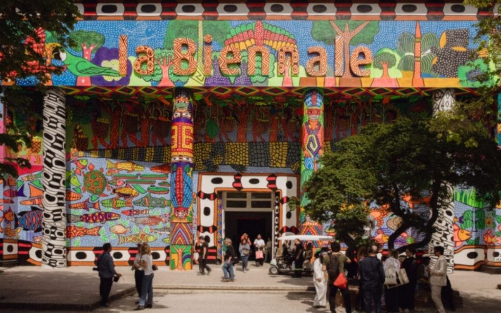 Główny Pawilon Biennale w Wenecji z wejściem pokrytym intensywnie kolorowym muralem w stylu sztuki ludowej. Zgromadzeni zwiedzający.