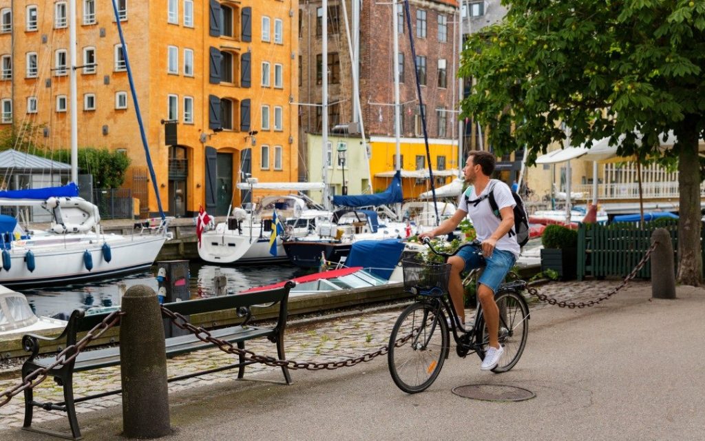 Kanał w Kopenhadze z zacumowanymi jachtami i kolorowymi kamienicami w tle, otoczony zielenią i ścieżką rowerową