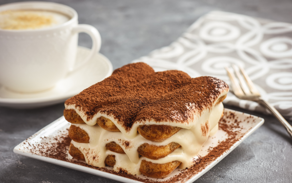 Klasyczne włoskie tiramisu, złożone z warstw biszkoptów i kremu, posypane kakao. Deser podany obok filiżanki cappuccino.