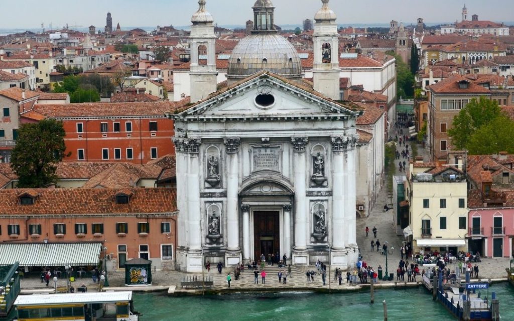 Kościół Redentore na wyspie Giudecca, widziany z góry. Biała, barokowa fasada nad kanałem, otoczona czerwonymi dachami.