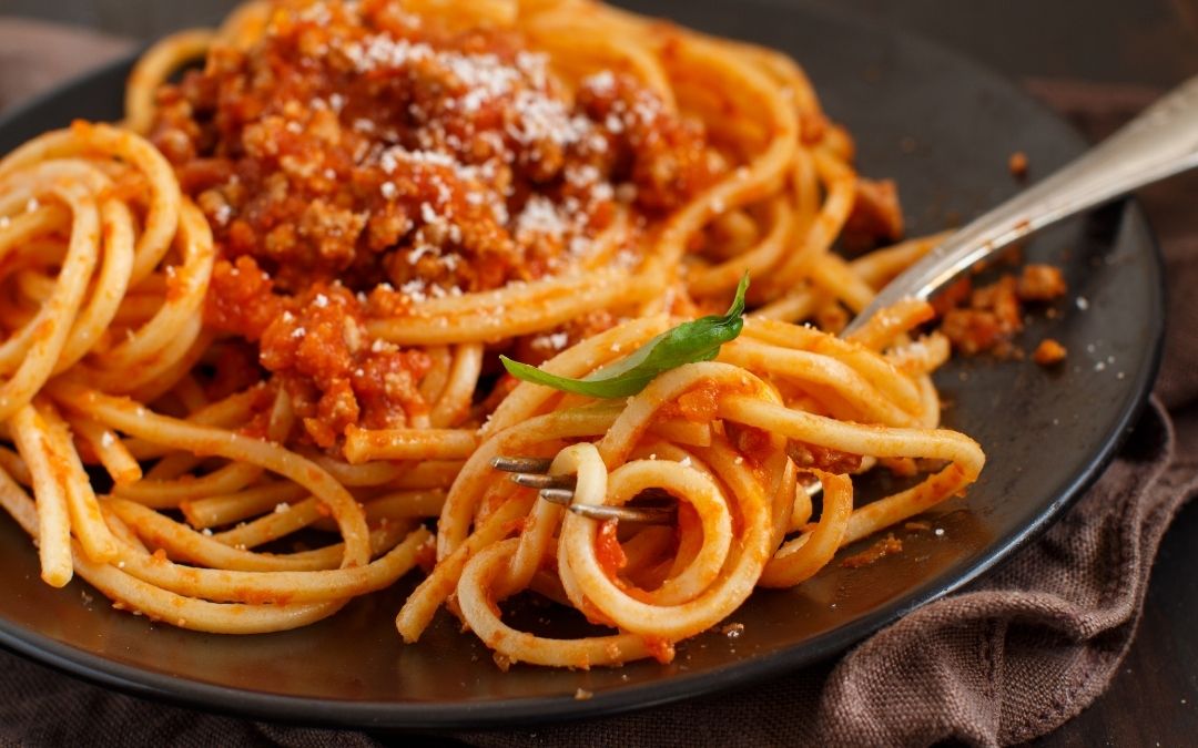 Makaron bucatini z sosem ragù, obficie posypany parmezanem. Widelec unosi porcję pasty. Bogate, zbliżone ujęcie.