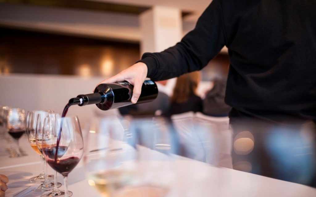 Mężczyzna nalewa czerwone wino z ciemnej butelki do kieliszka. Zbliżenie na degustację wina, na długim, białym stole.