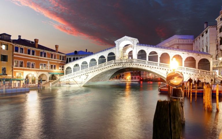 Most Rialto w Wenecji o zmierzchu. Biały łuk oświetlony lampami, odbija się w Canal Grande. Nad nim dramatyczne, czerwone niebo.