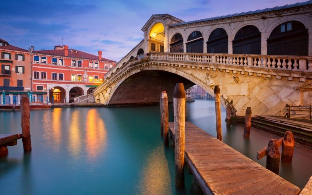 Most Rialto w Wenecji o zmierzchu. Oświetlony kamienny łuk odbija się w spokojnym Canal Grande, z drewnianym pomostem na pierwszym planie.
