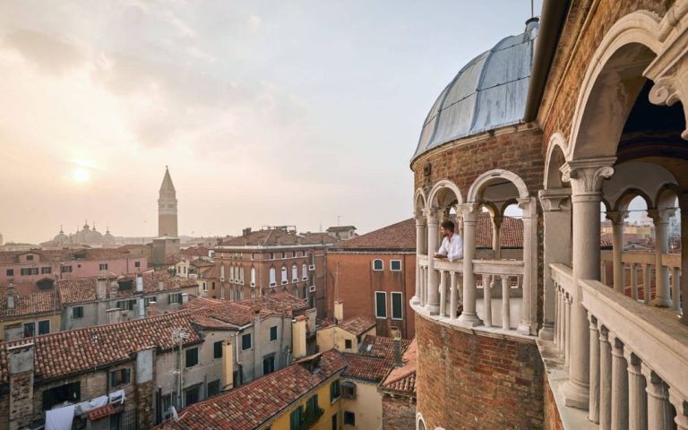 Panoramiczny widok na dachy Wenecji z tarasu pałacu. W tle dzwonnica św. Marka, w prawym kadrze osoba na arkadowym balkonie.