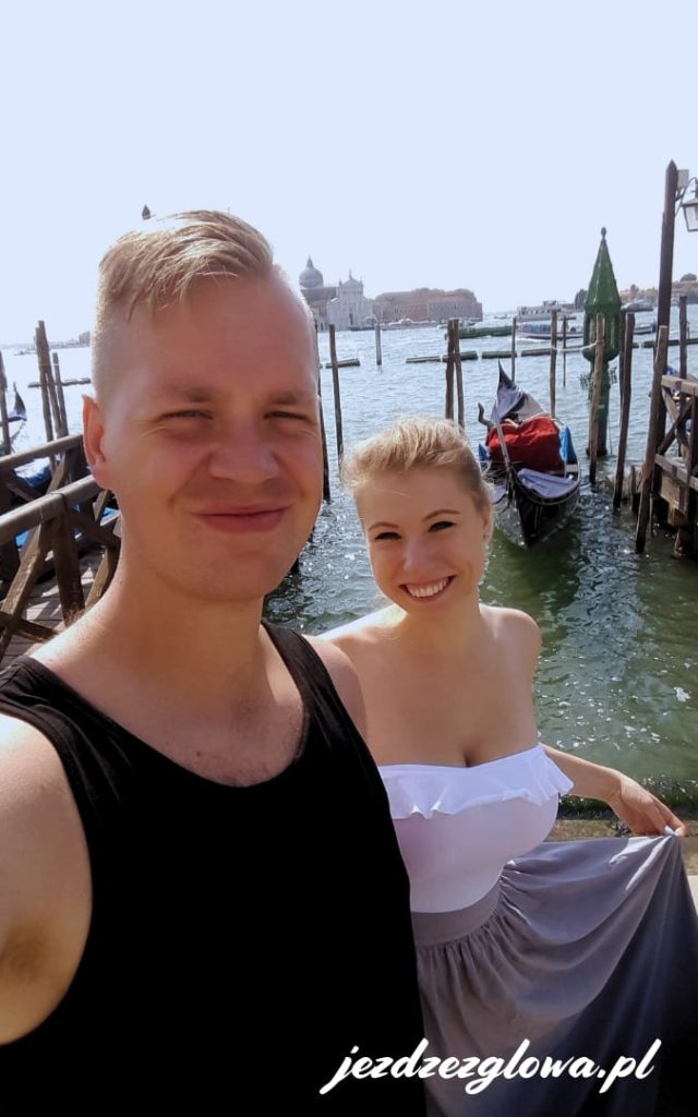Para turystów robiąca selfie przy przystani gondoli w Wenecji z laguną w tle