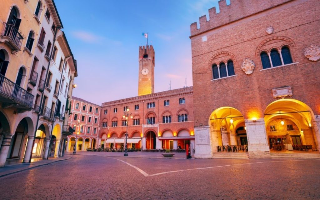 Piazza dei Signori w Treviso o zmierzchu. Ceglana wieża zegarowa góruje nad placem otoczonym arkadowymi pałacami.