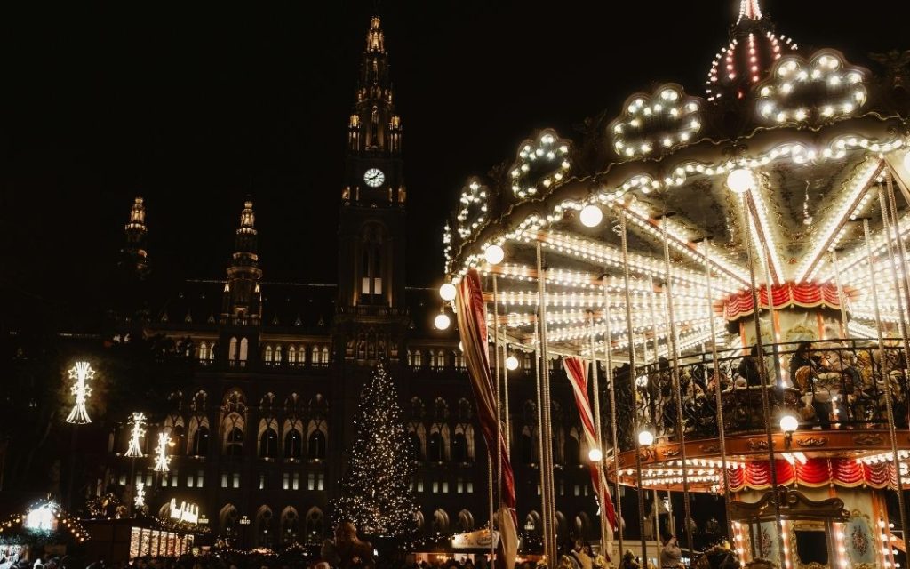 Rynek świąteczny nocą. Wiedeński Ratusz i choinka w tle, a na pierwszym planie jasna, rozświetlona karuzela.