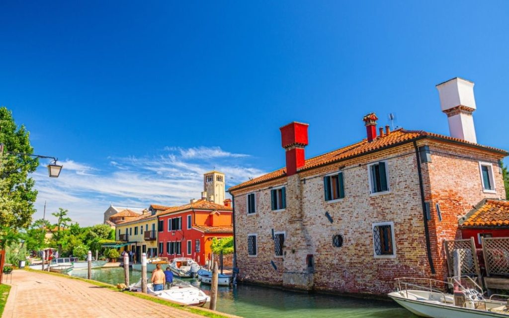 Słoneczny kanał na wyspie Torcello (Laguna Wenecka). Ceglane domy z czerwonymi akcentami i wyraźna wieża w tle.