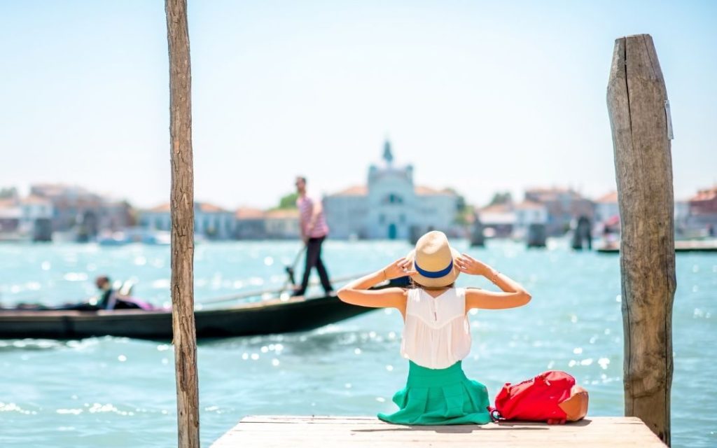 Turystka w kapeluszu i zielonej spódnicy siedzi na drewnianym pomoście w Wenecji. Podziwia widok na lagunę, obok przepływa gondola.
