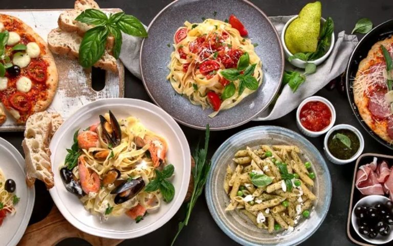 Ujęcie z góry bogaty wybór włoskich potraw, w tym pizze, trzy rodzaje past (seafood, pesto, pomidorowa), szynka parmeńska i bazylia.