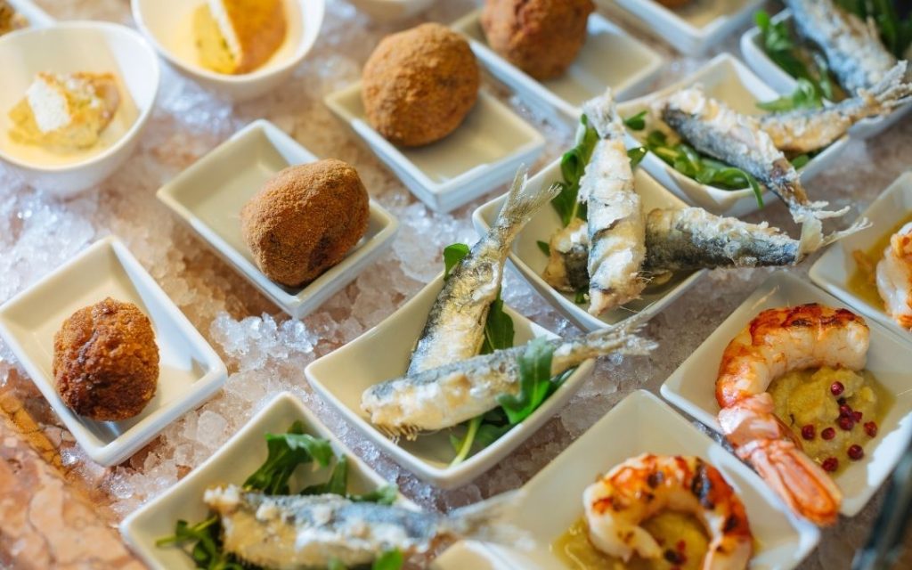 Weneckie przekąski cicchetti. Różnorodne małe dania, w tym smażone ryby i krewetki, podane na białych talerzykach na lodzie