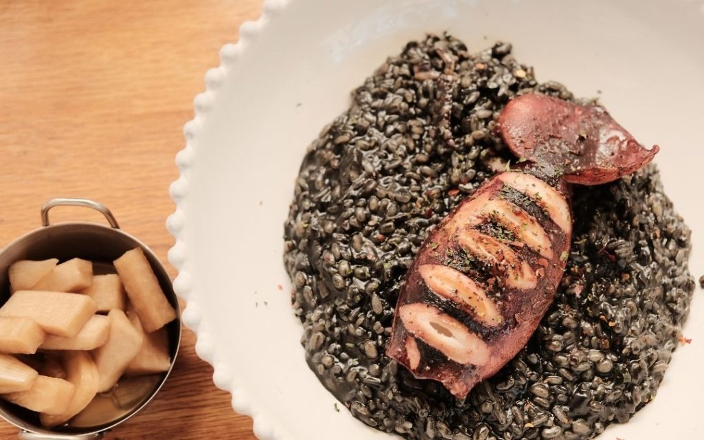 Weneckie risotto al nero di seppia (czarny ryż) z grillowanym kalmarem. Danie podane w białej misce, obok mała miseczka pikli.