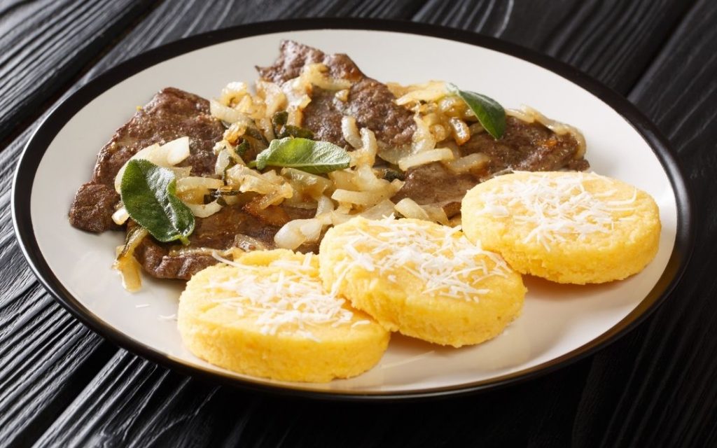 Włoskie danie wątróbka (Fegato) z cebulą i polenta. Trzy okrągłe porcje polenty posypane serem na białym talerzu.