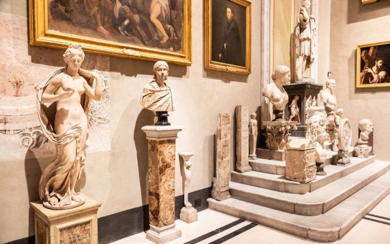 Wnętrze muzeum w Wenecji. Ekspozycja marmurowych rzeźb i popiersi na stopniach, z obrazami w złotych ramach wiszącymi w tle.
