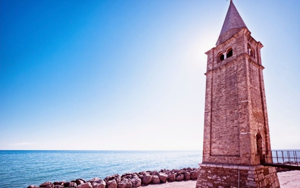 Wysoka, ceglana dzwonnica w Caorle nad Morzem Adriatyckim. Charakterystyczna stożkowa wieża stoi na brzegu, przy falochronie z kamieni.