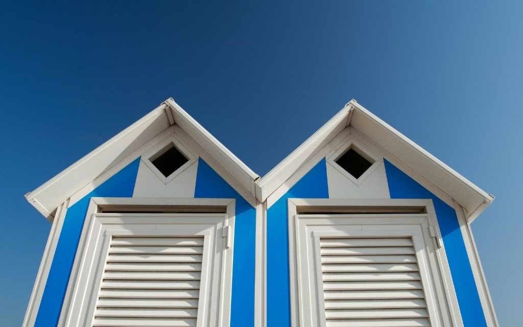 Zbliżenie na dwie sąsiadujące kabiny plażowe w Bibione. Biało-niebieskie pasiaste ściany i lamelowe drzwi pod jasnym, bezchmurnym niebem.
