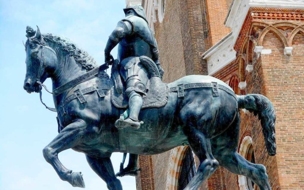 Zbliżenie na pomnik konny Bartolomeo Colleoniego w Wenecji. Jeździec w zbroi, na dynamicznym koniu, na tle ceglanej architektury.