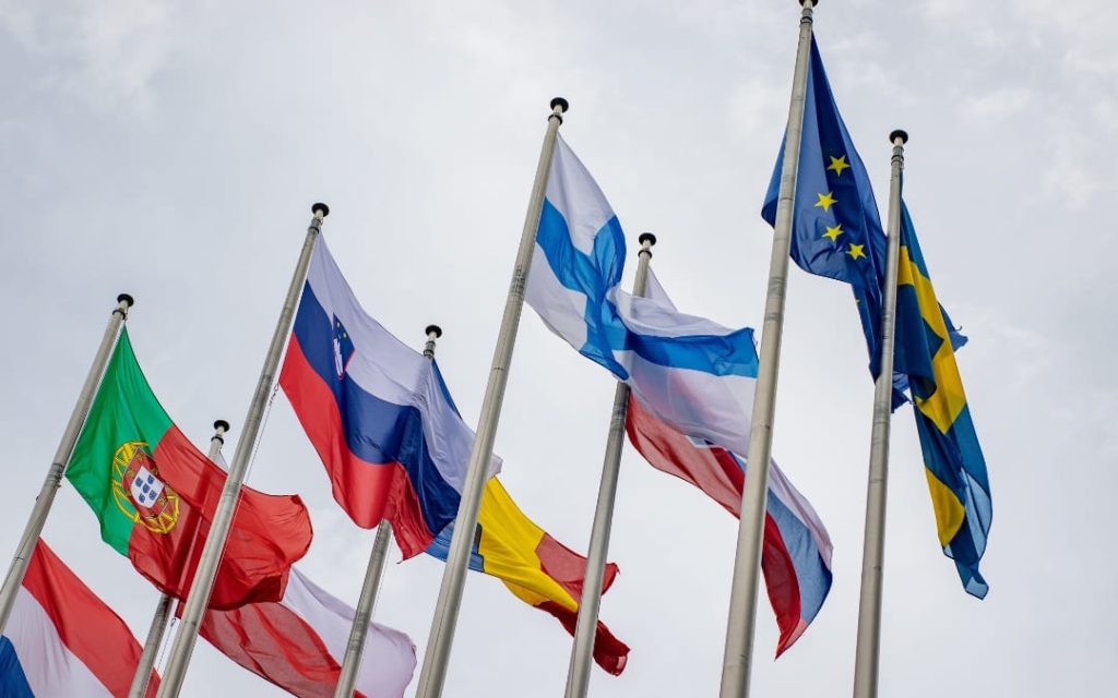 Flagi państw członkowskich Unii Europejskiej powiewające na masztach na tle zachmurzonego nieba