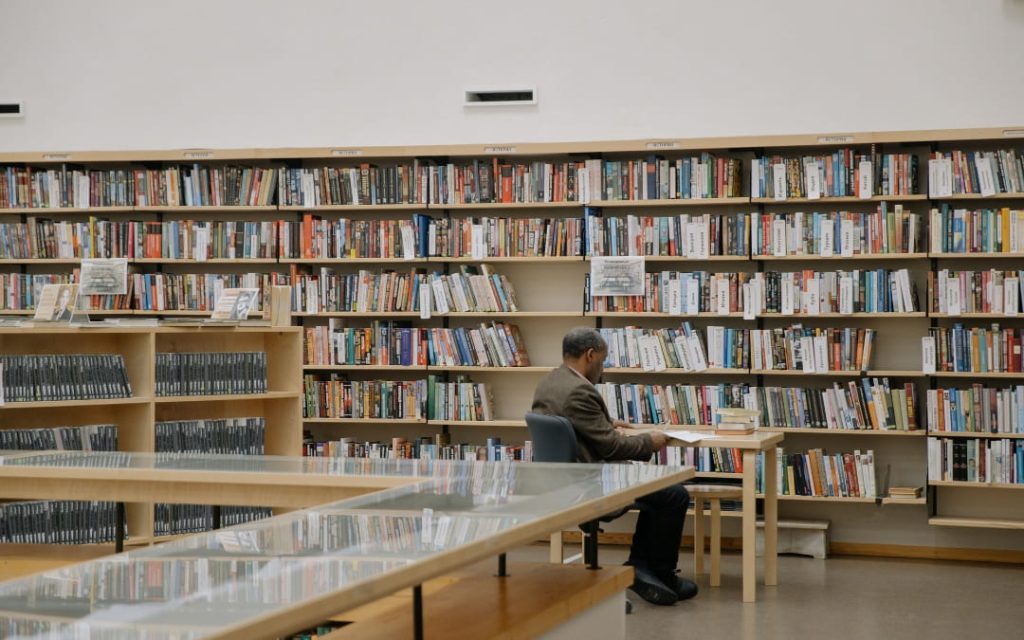 Mężczyzna czytający książkę przy stoliku w bibliotece z wypełnionymi regałami w tle
