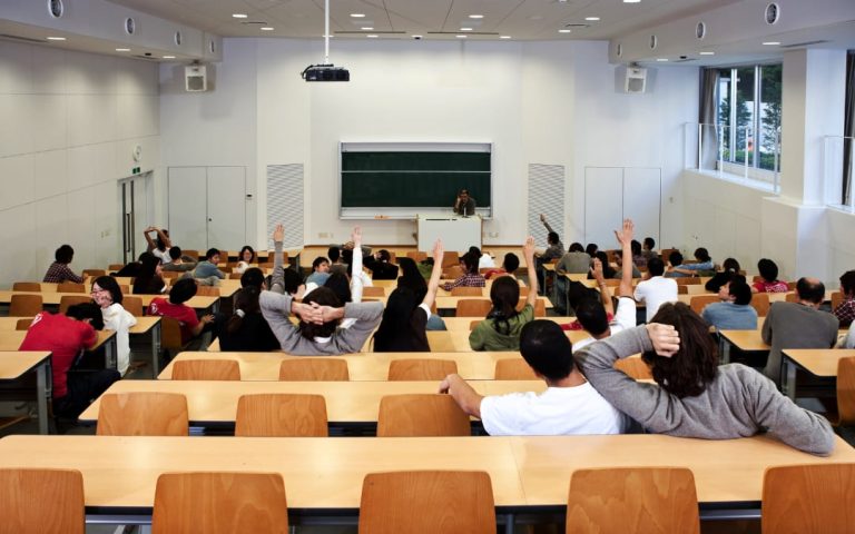 Studenci siedzący w nowoczesnej sali wykładowej z podniesionymi rękami podczas zajęć na uczelni
