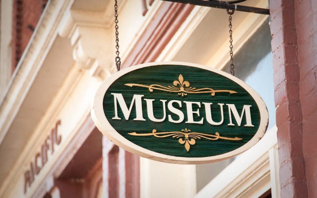 Zielona owalna tablica z napisem Museum i złotymi ornamentami wisząca na łańcuchu przy budynku