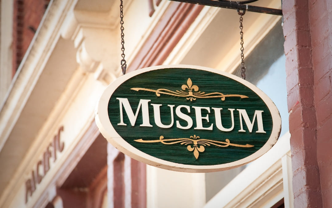 Zielona owalna tablica z napisem Museum i złotymi ornamentami wisząca na łańcuchu przy budynku
