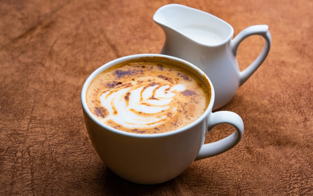 Biała filiżanka kawy z latte art i dzbanuszkiem mleka na brązowym tle