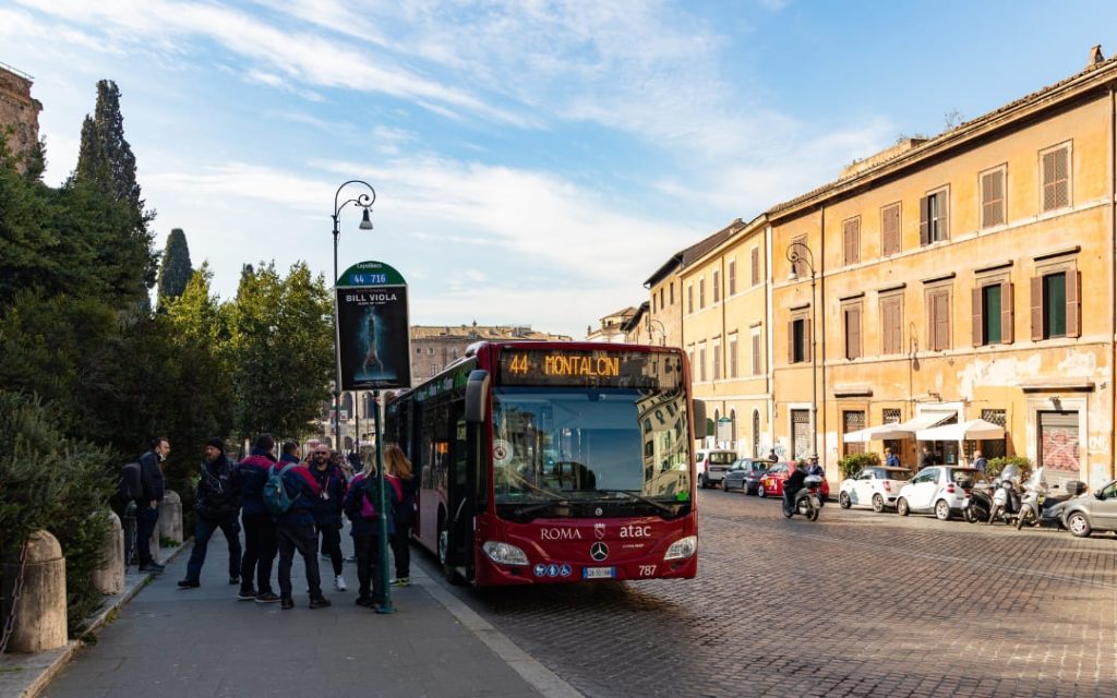 Czerwony autobus ATAC linii 44 jadący w stronę Montalcini na tle kamienic przy brukowanej rzymskiej ulicy