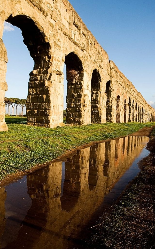 Długi ciąg kamiennych łuków akweduktu Aqua Claudia odbijający się w kałuży na trawiastej ścieżce