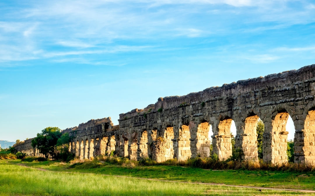 Długi ciąg wysokich, kamiennych łuków rzymskiego akweduktu Aqua Claudia na tle zielonej trawy i błękitnego nieba