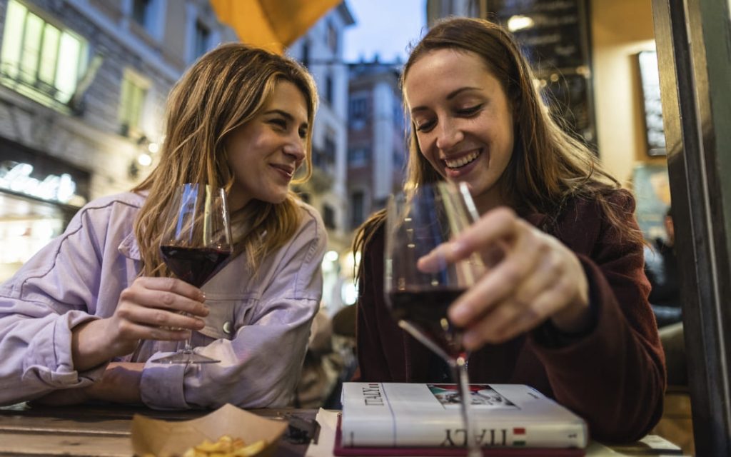 Dwie uśmiechnięte kobiety pijące czerwone wino przy kawiarnianym stoliku w rzymskiej dzielnicy o zmierzchu