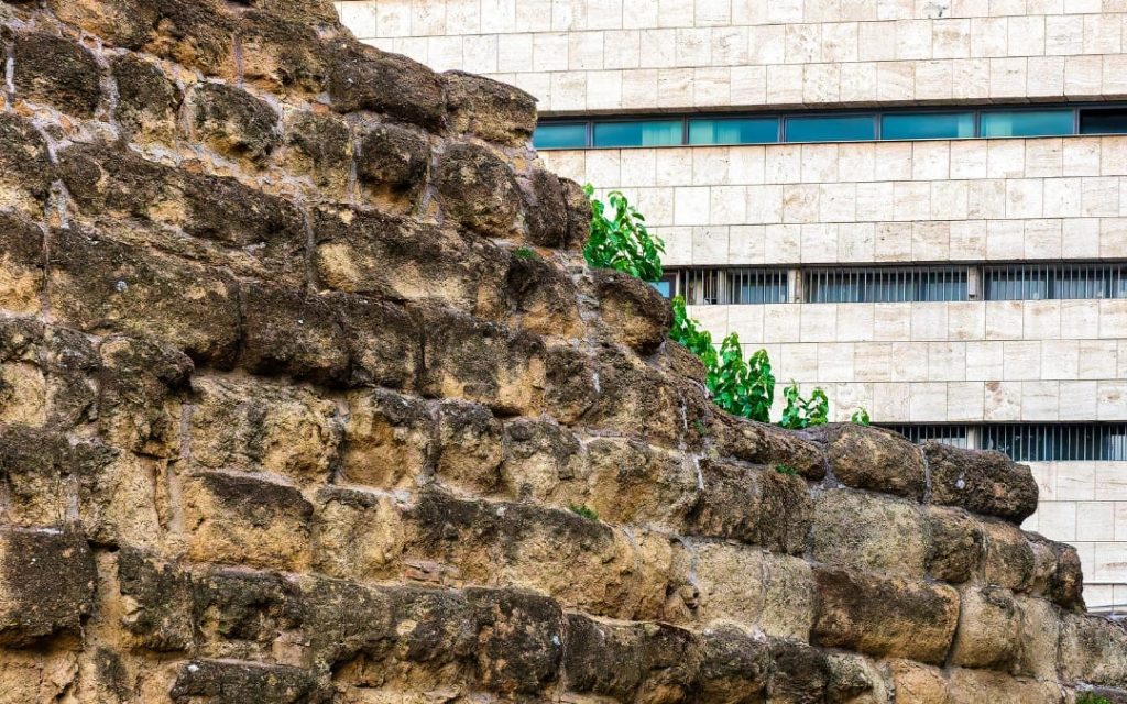 Fragment starożytnego Muru Serwiańskiego z ciemnych bloków tufu na tle nowoczesnego, jasnego biurowca
