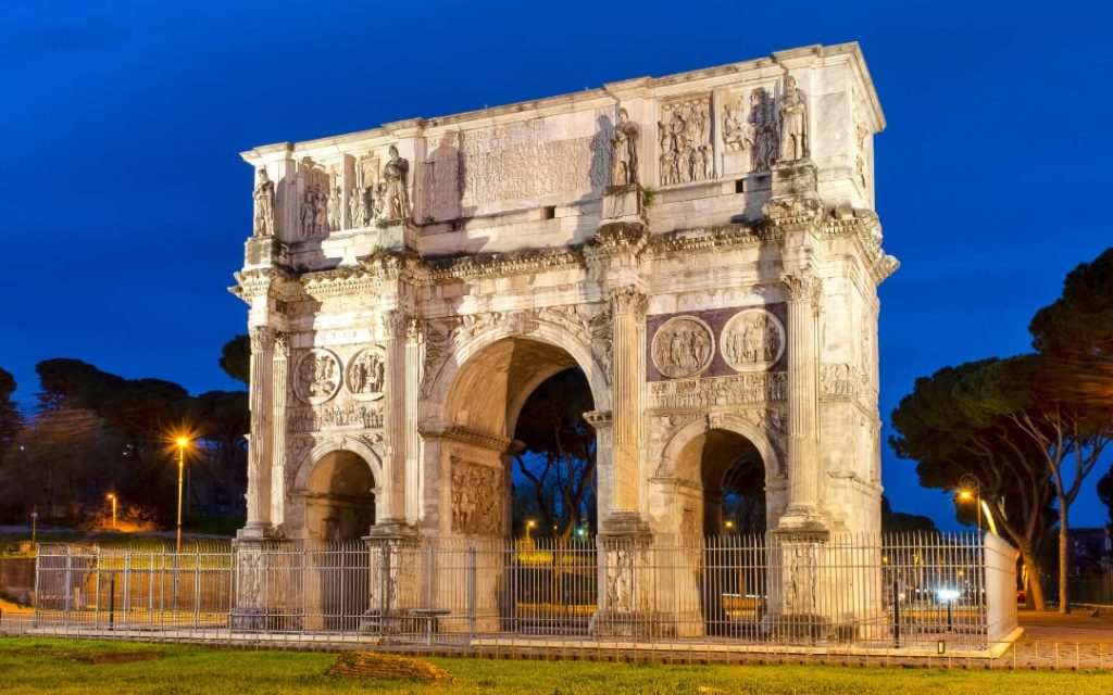 Oświetlony nocą trójprzęsłowy Łuk Konstantyna Wielkiego za metalowym ogrodzeniem w Rzymie