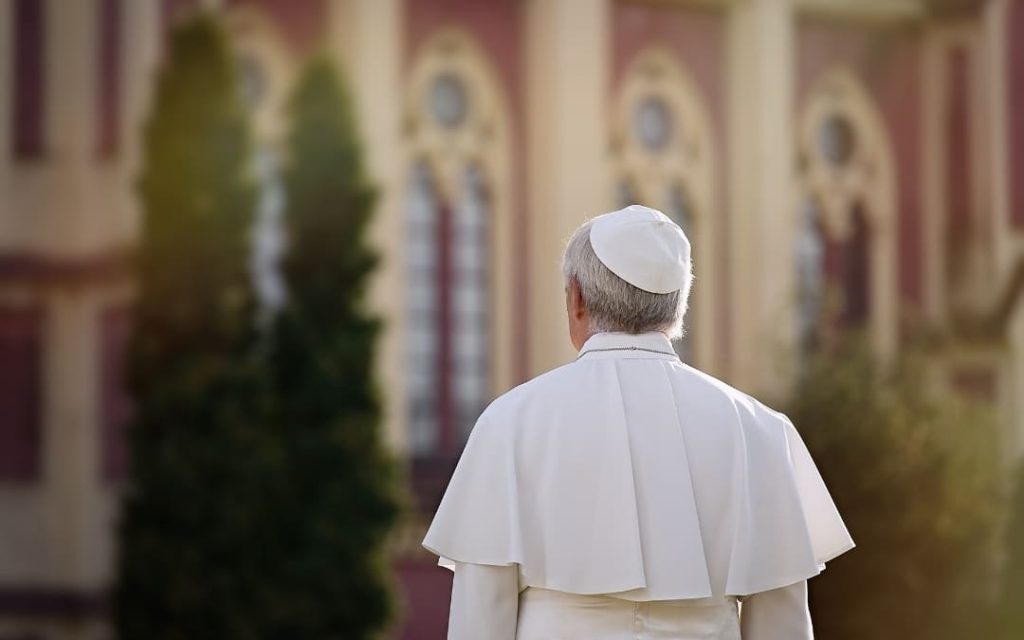 Papież w białej sutannie, widziany od tyłu na tle neogotyckiej fasady budynku