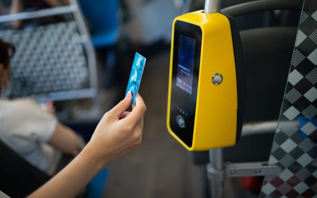 Pasażerka przykładająca niebieską kartę płatniczą do żółtego kasownika zbliżeniowego w autobusie