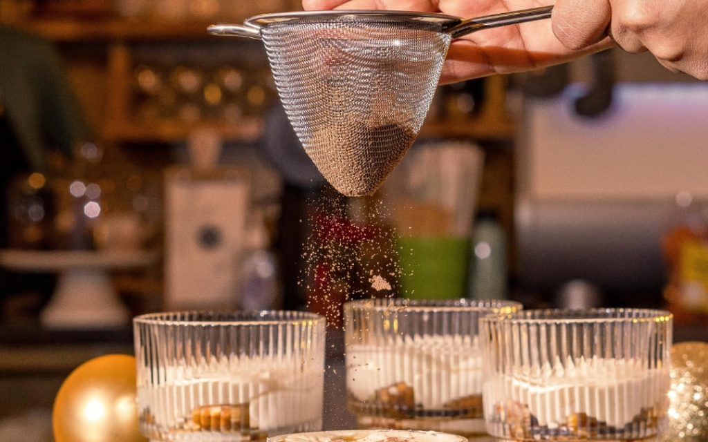 Przesiewanie ciemnego kakao przez sitko na desery tiramisu w szklanych naczyniach