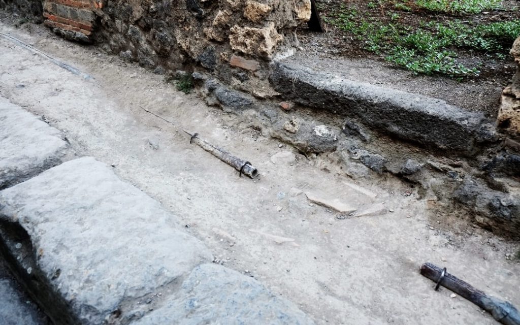 Starożytne, ołowiane rury wodociągowe wyłaniające się z piaszczystego gruntu obok kamiennego muru w Pompejach