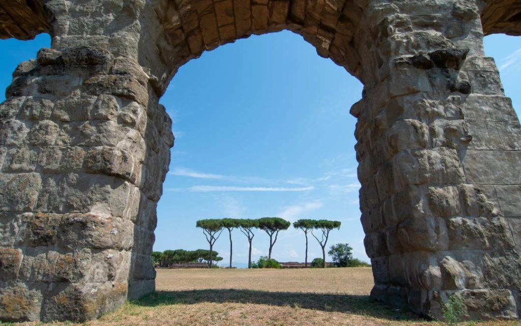 Widok przez wysoki, kamienny łuk rzymskiego akweduktu na rząd pinii rosnących na otwartym polu